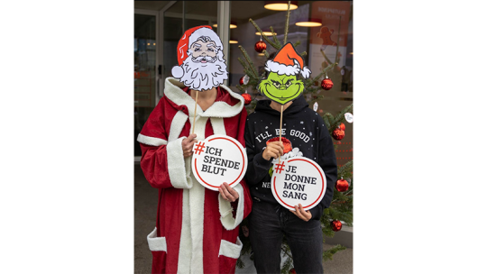 Weihnachtsmann und Grinch vor Tannenbaum mit #ichspendeblut Schild